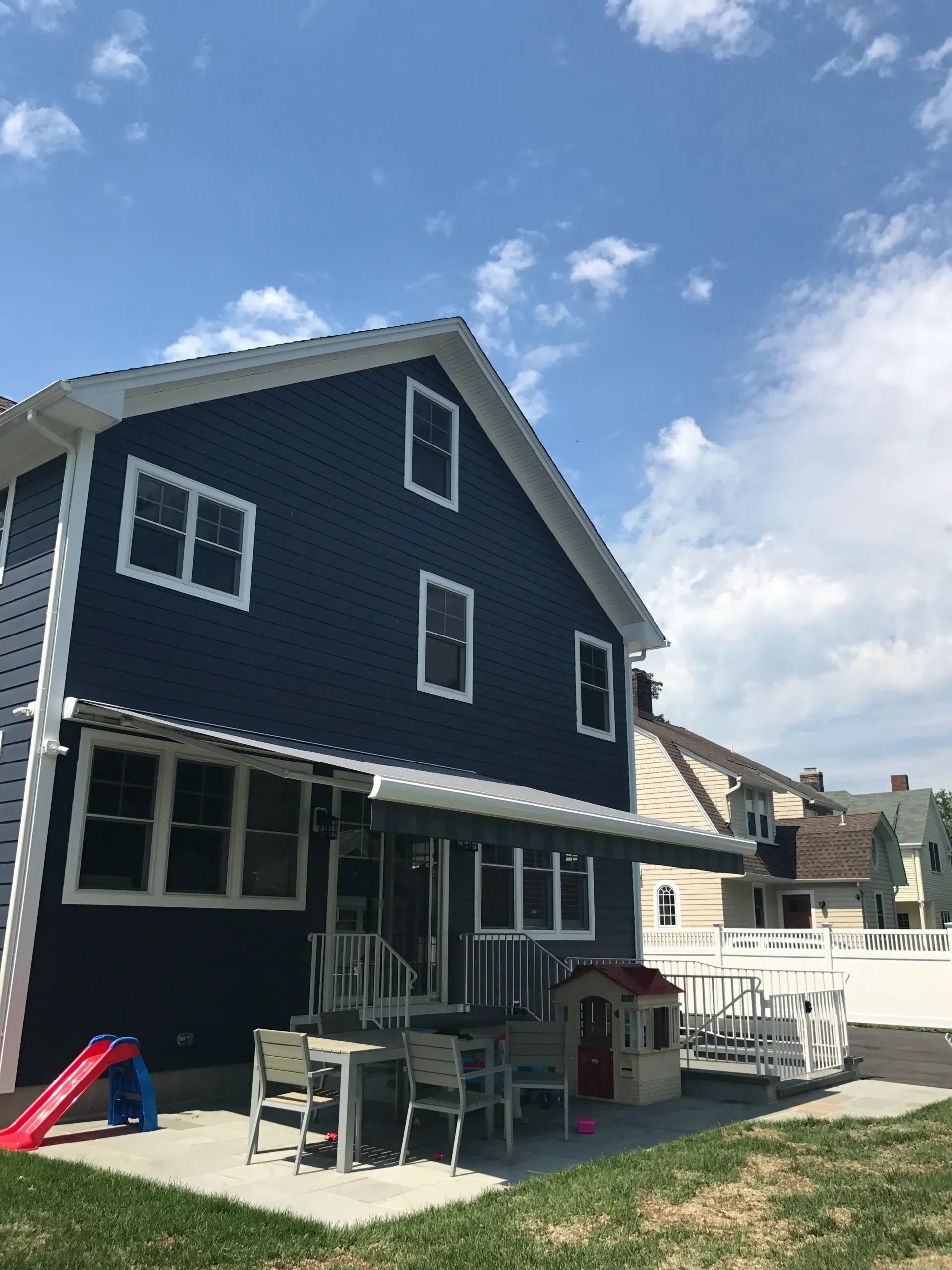 Retractable awning extended over a backyard patio of a dark-colored home with outdoor seating and a fenced yard.