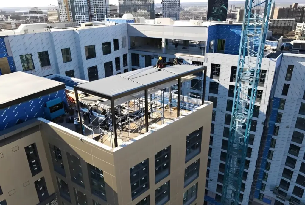 Construction workers install a steel frame on a partially completed rooftop terrace, surrounded by a high-rise urban skyline.