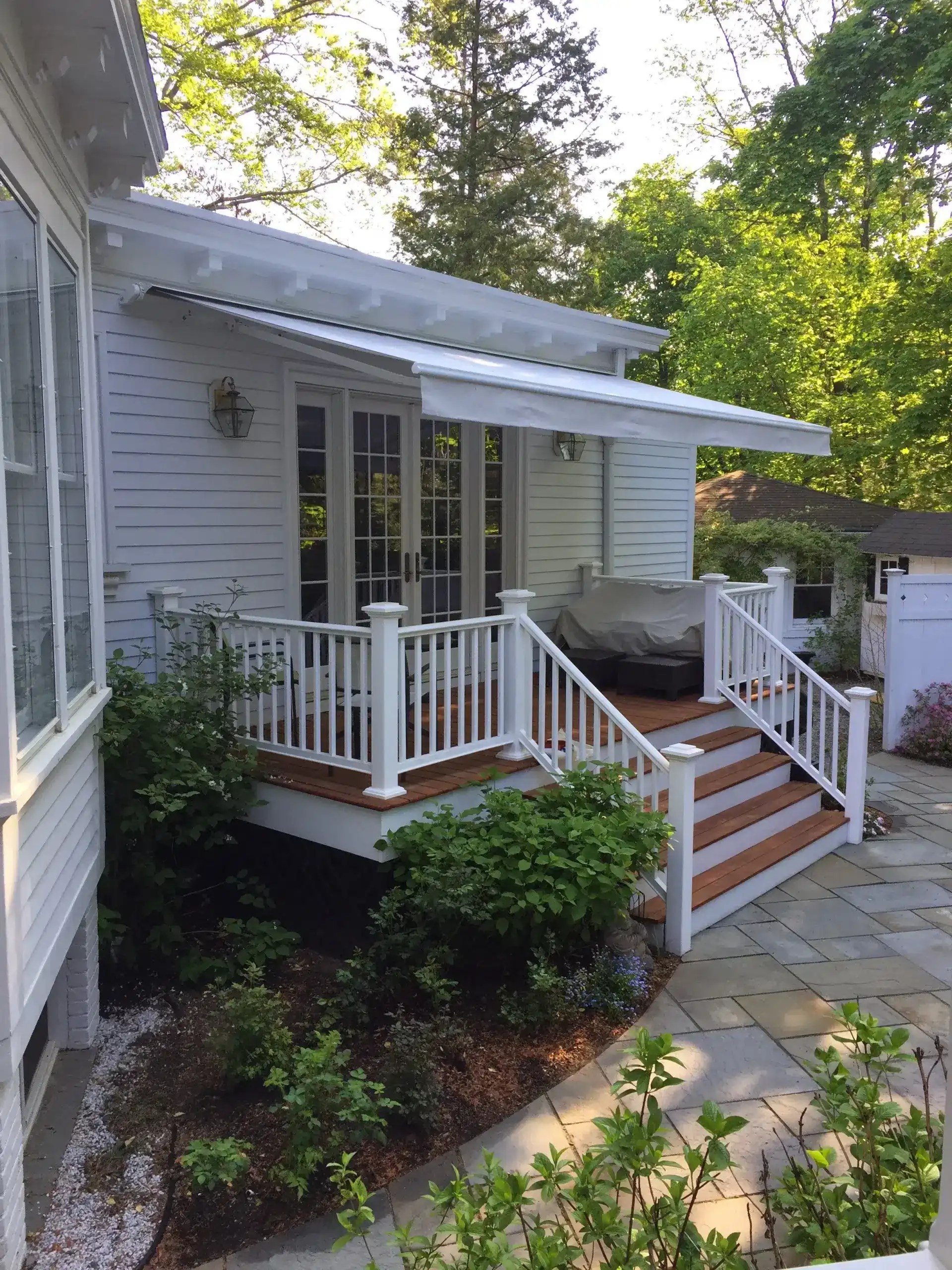 Retractable awning extended over a raised porch with white railings, French doors, and surrounding garden landscaping.